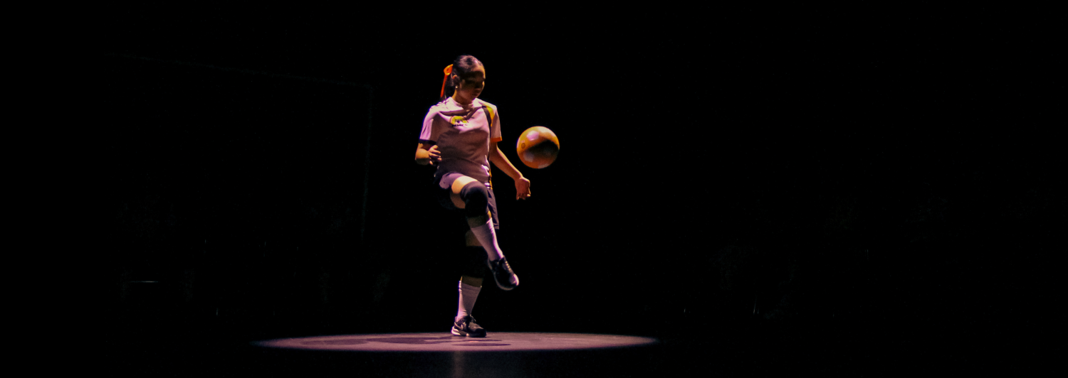 A young woman on a stage wearing a soccer uniform, kicking a soccer ball in the air